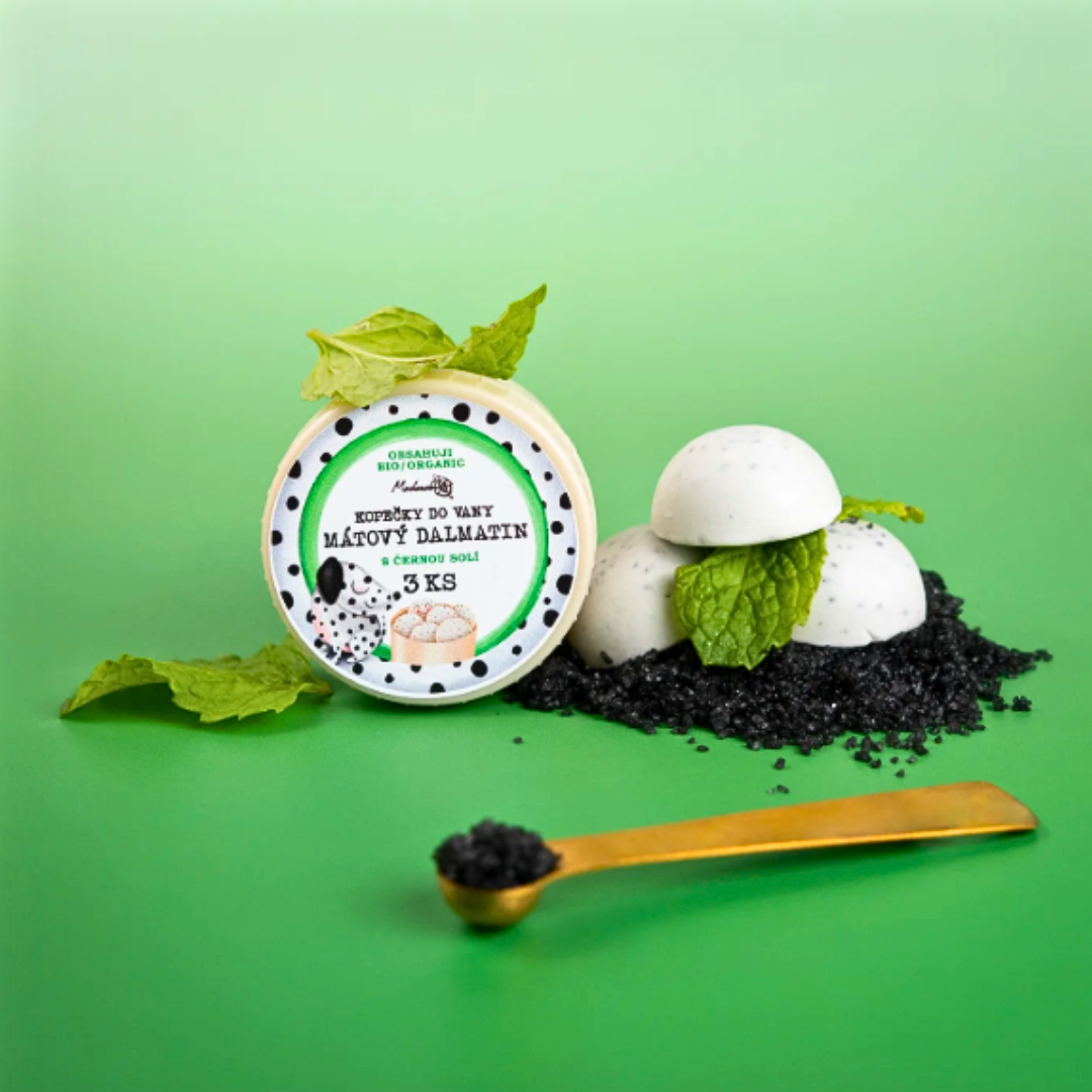 Mint “Dalmatian” bath melts with black lava salt – open tin (3 pcs) surrounded by white speckled bath domes, mint leaves and a spoonful of black salt on a green background.